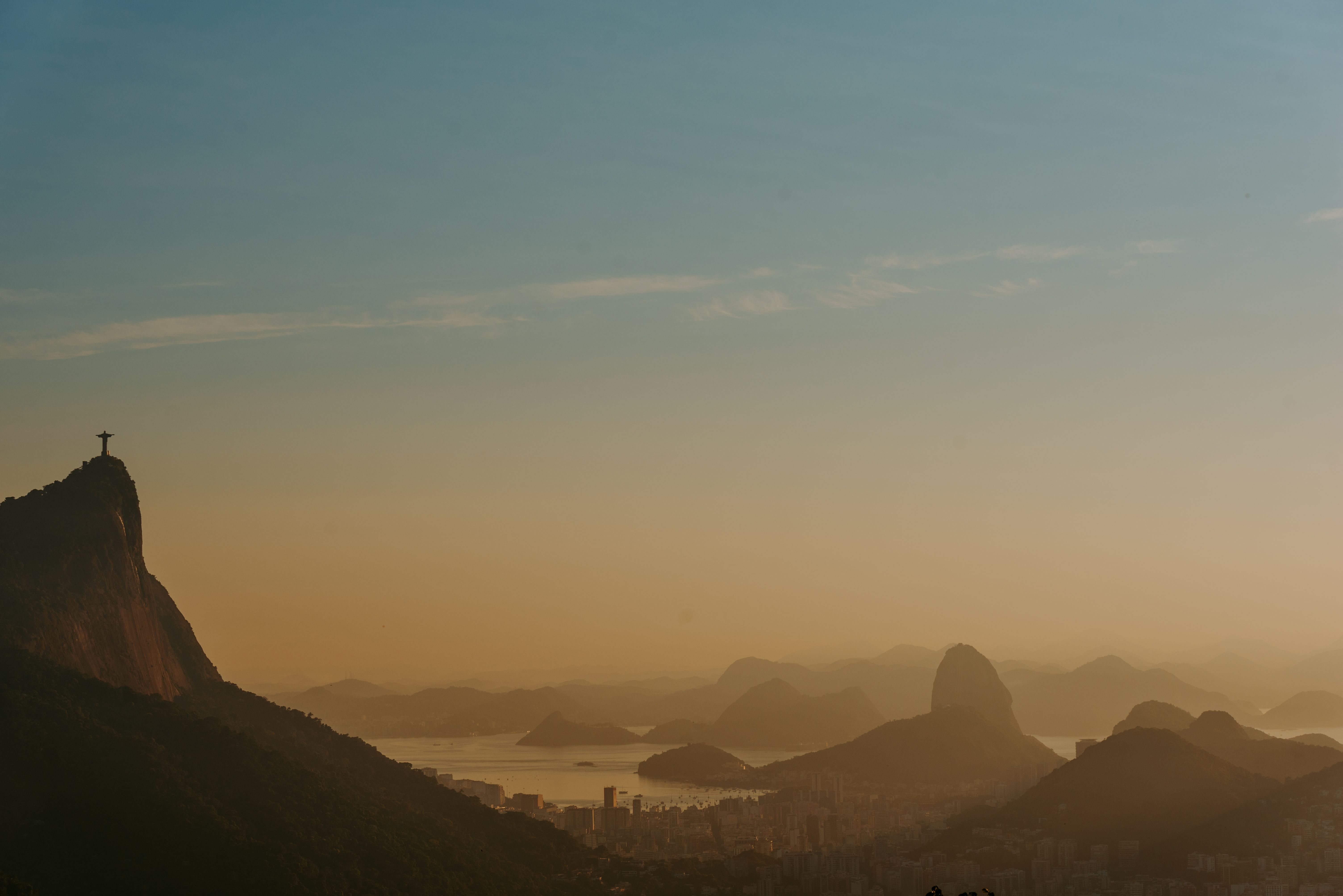 Christ the Redeemer statue bathed in warm golden light at sunset, with Rio de Janeiro's urban sprawl and Guanabara Bay stretching below