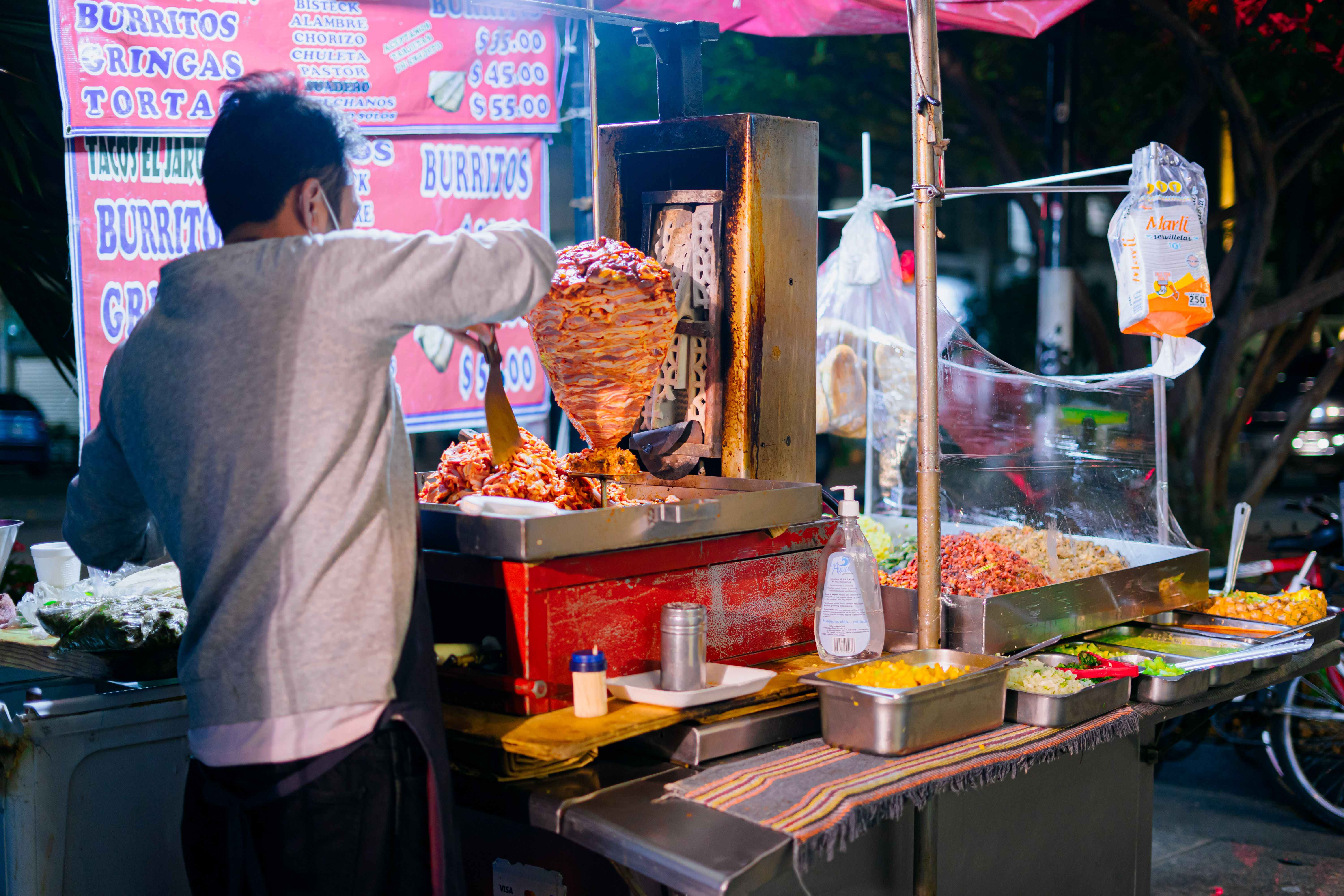 A street vendor slicing tacos al pastor from a vertical spit with pineapple on top at a busy Mexico City taquería at night