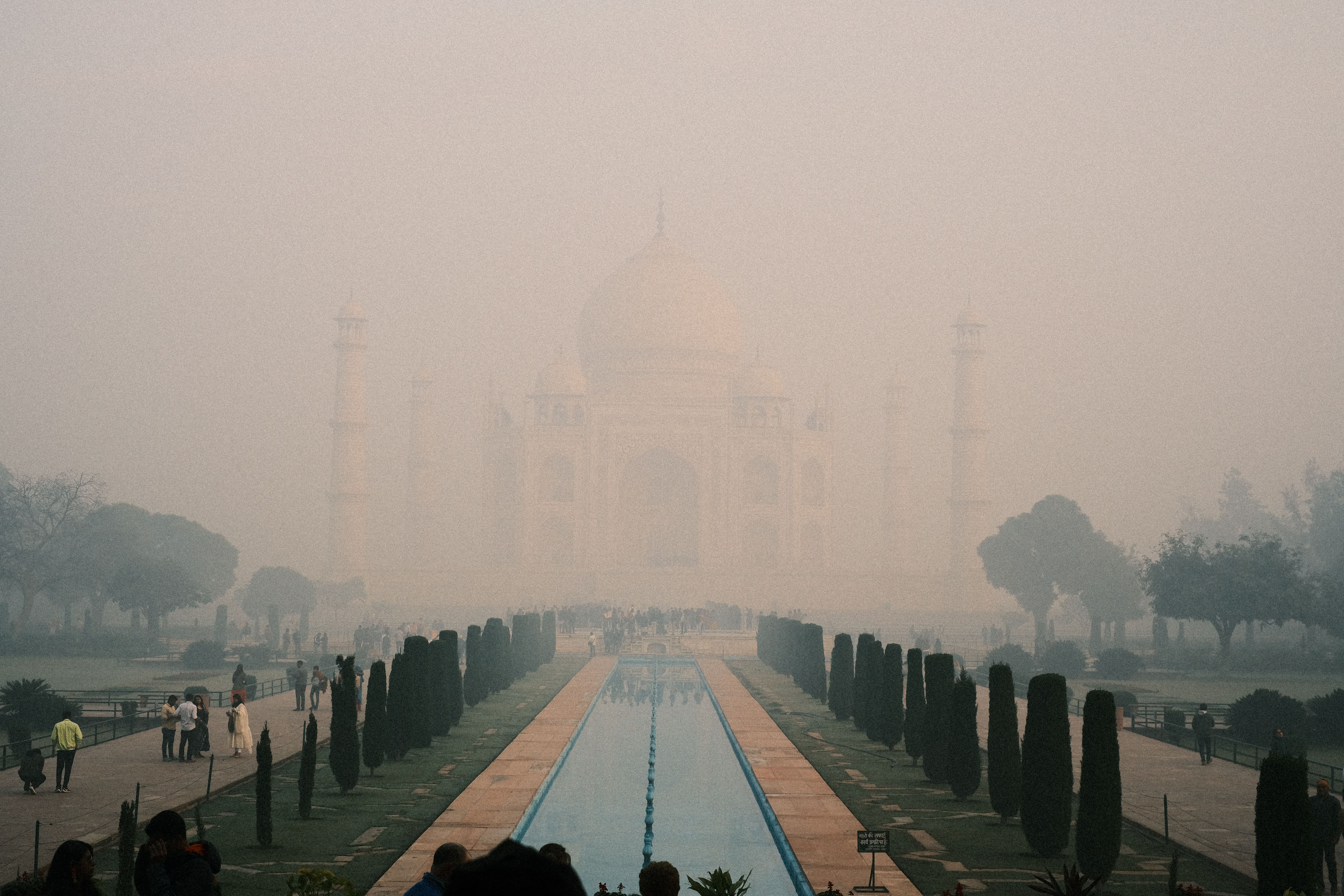 The Taj Mahal bathed in soft golden dawn light with morning mist rising from the Yamuna River, its white marble dome and minarets reflected in the long ornamental pool of the main approach