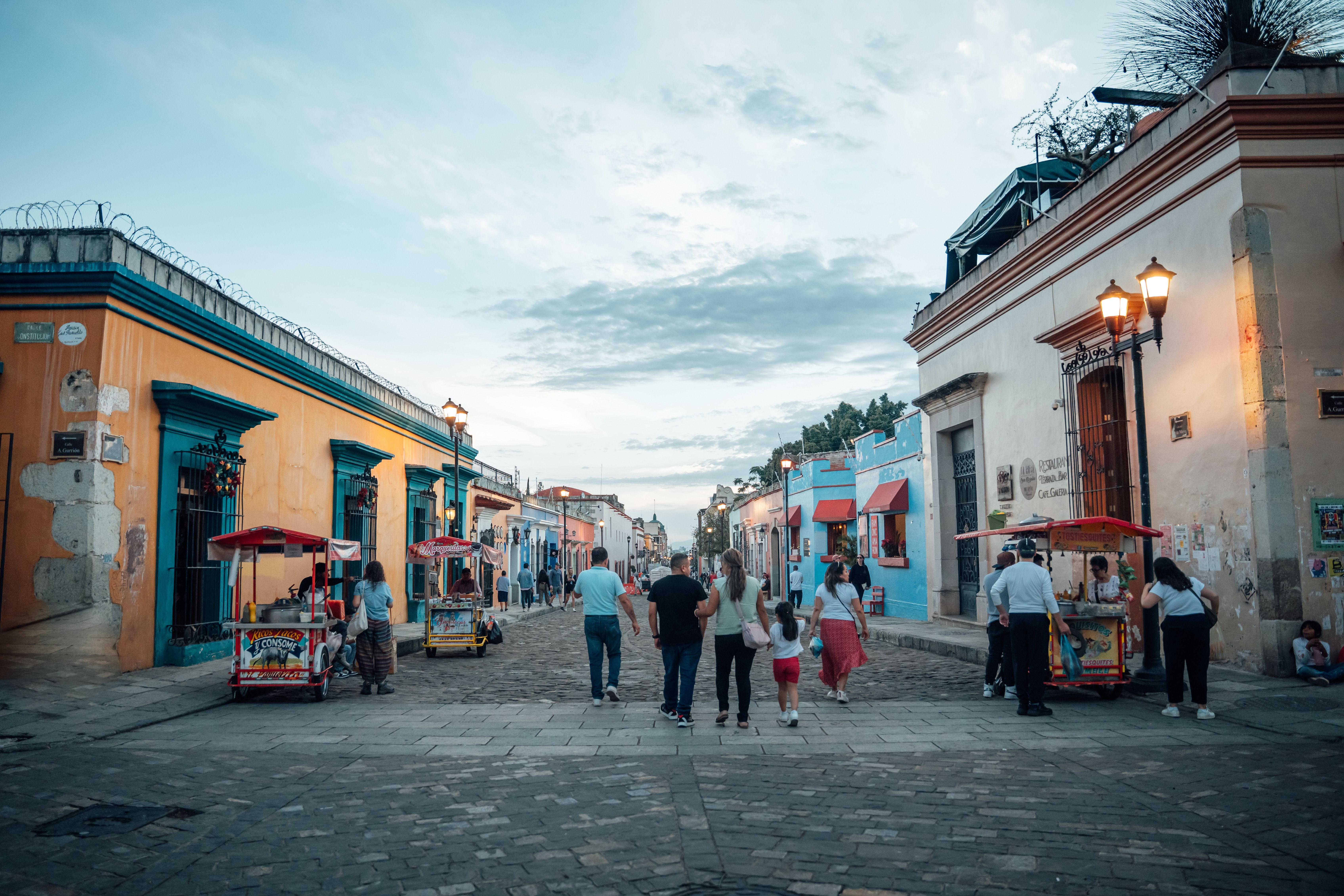Colorful cobblestone street in Oaxaca de Juárez leading toward the ornate Baroque facade of the Santo Domingo church, with vendors and flowering trees lining the sidewalk
