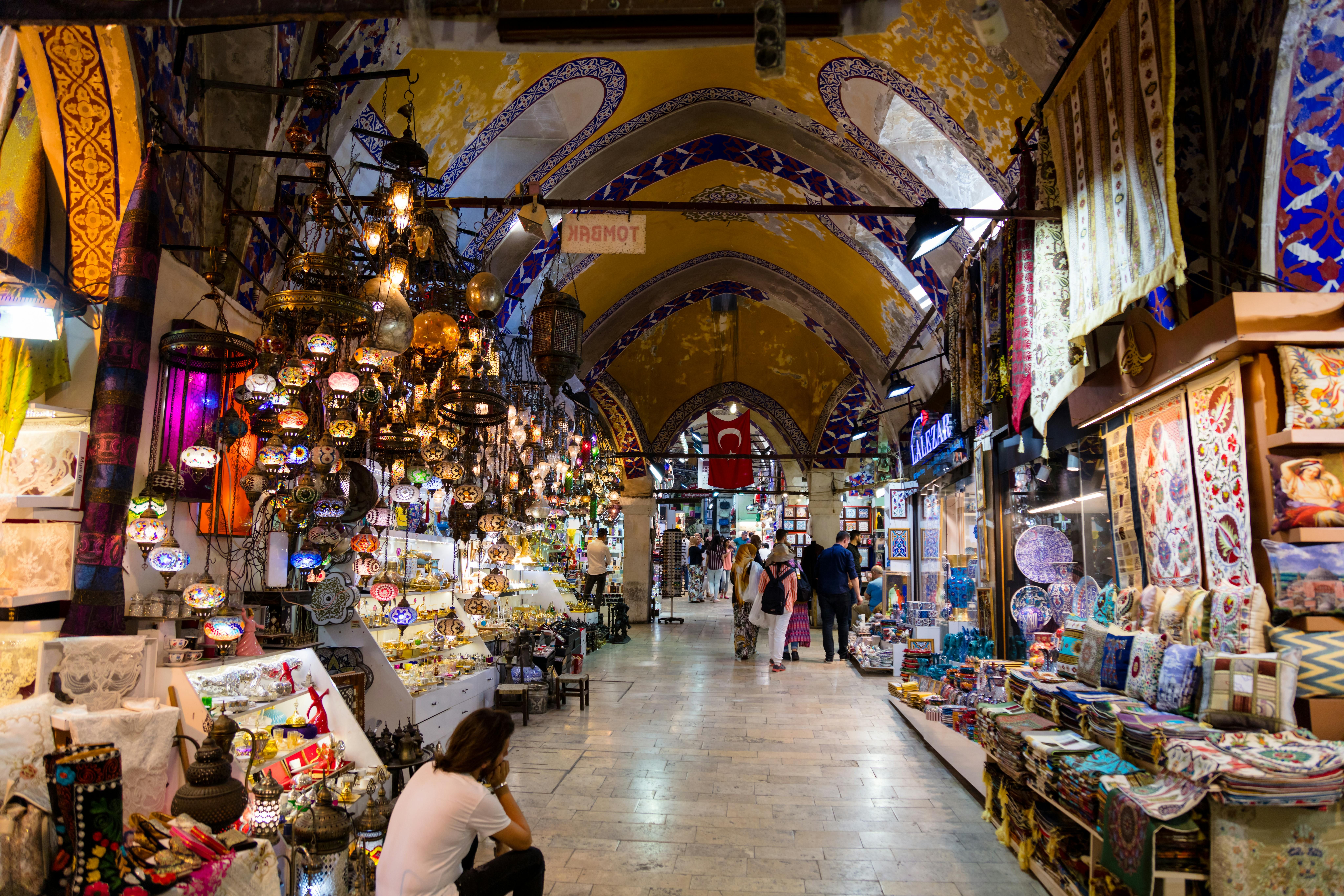 Ornate arched corridors of Istanbul's Grand Bazaar lined with colourful lanterns, textiles, and ceramics, with vendors and visitors moving through the golden-lit passageways