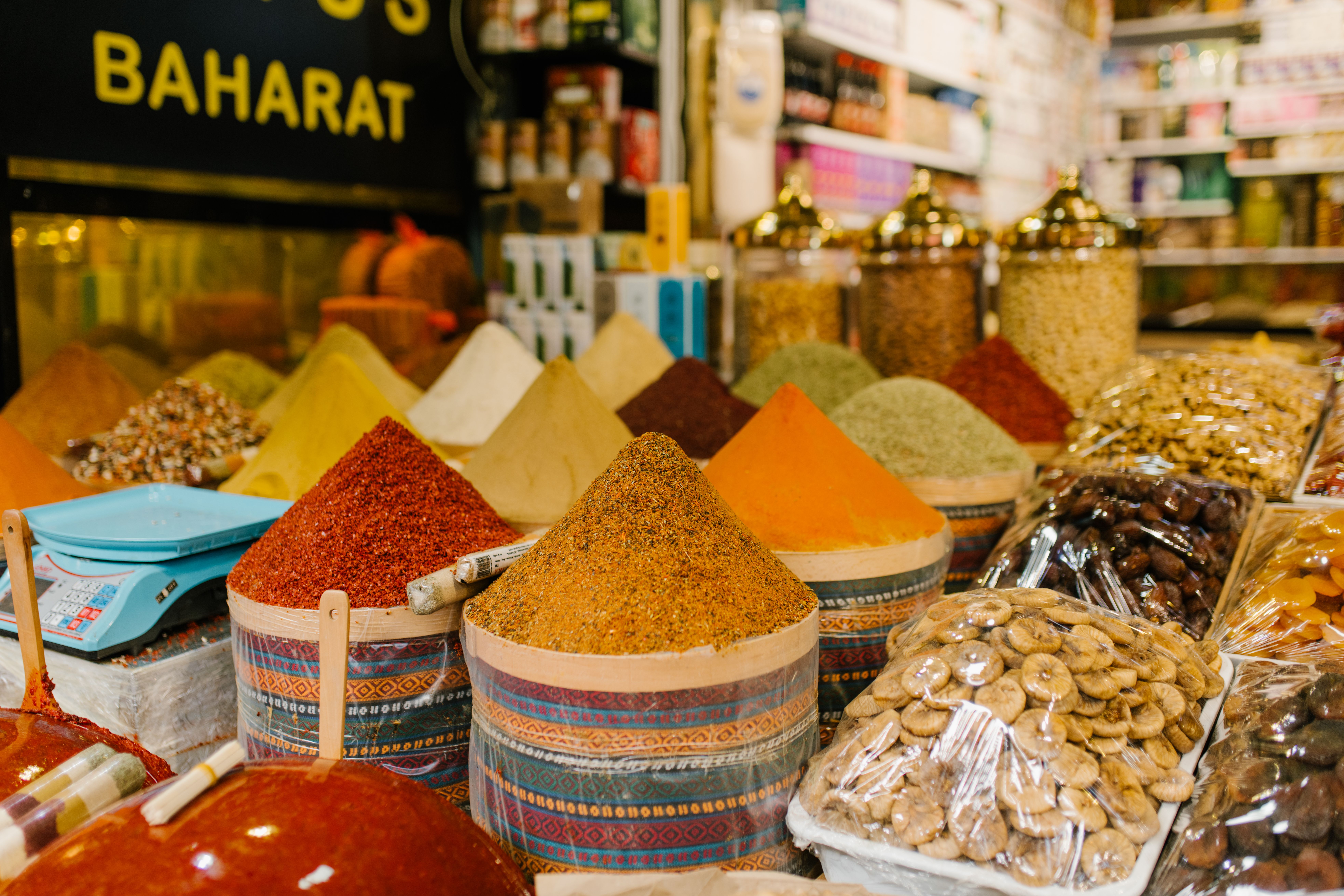 A spice market vendor holding a small pile of deep red saffron threads above a display of spices at a market stall, demonstrating quality to a prospective buyer